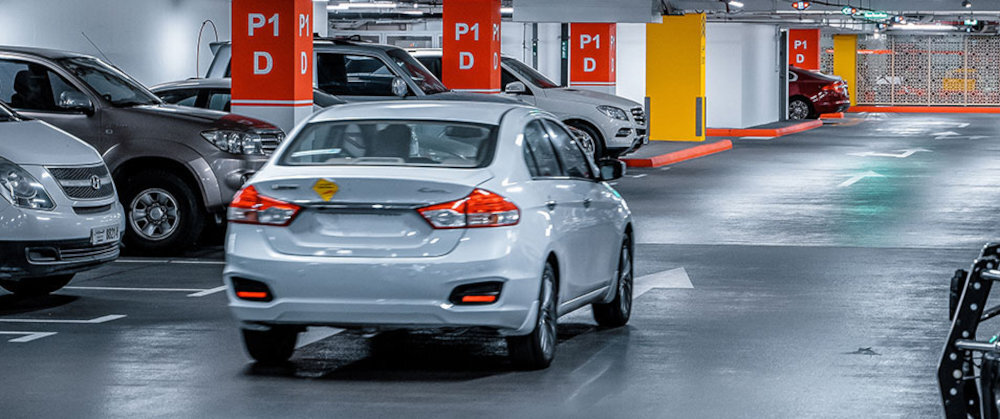 Samochód jedzie przez podziemny parking z oznaczeniami P1 D na kolumnach. W tle widoczne są zaparkowane samochody i oświetlenie sufitowe.
