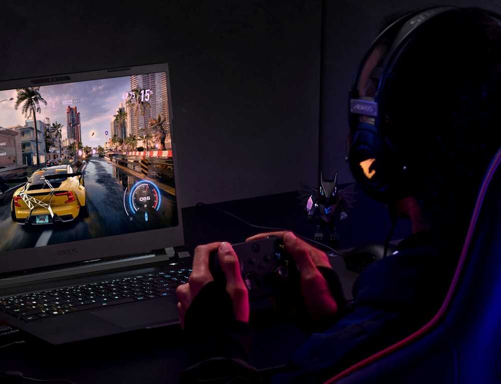 Osoba gra na laptopie, na ekranie widoczna jest gra wyścigowa z żółtym samochodem. Laptop ma podświetlaną klawiaturę i logo AORUS na obudowie.