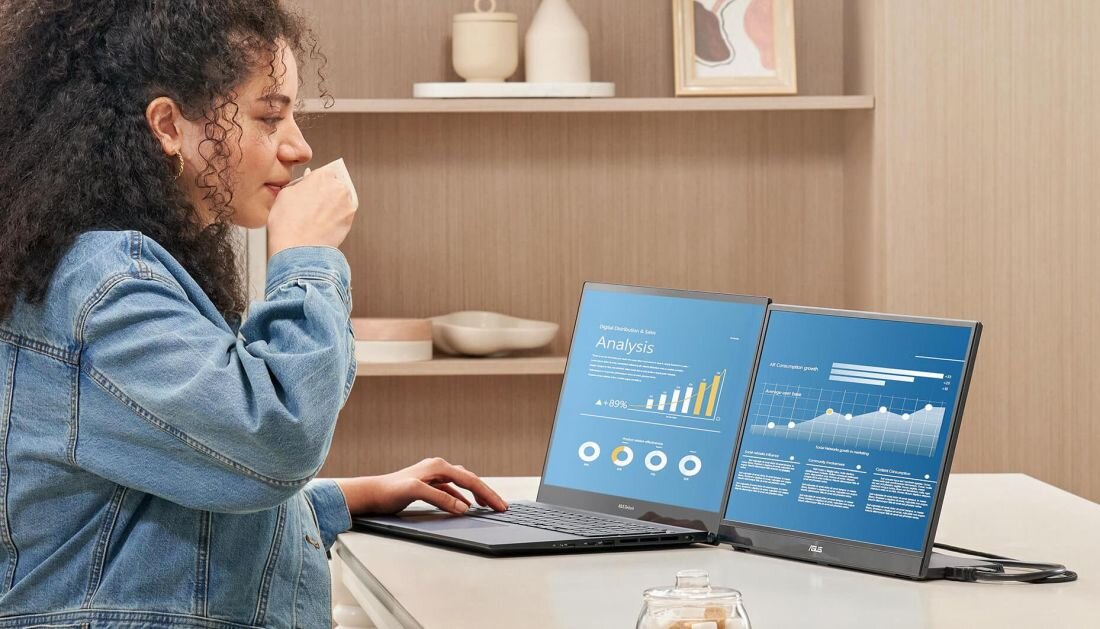Osoba siedzi przy stole, korzystając z laptopa i podłączonego monitora ASUS ZenScreen, na którym wyświetlane są wykresy i dane analityczne. W tle widać półkę z dekoracjami.