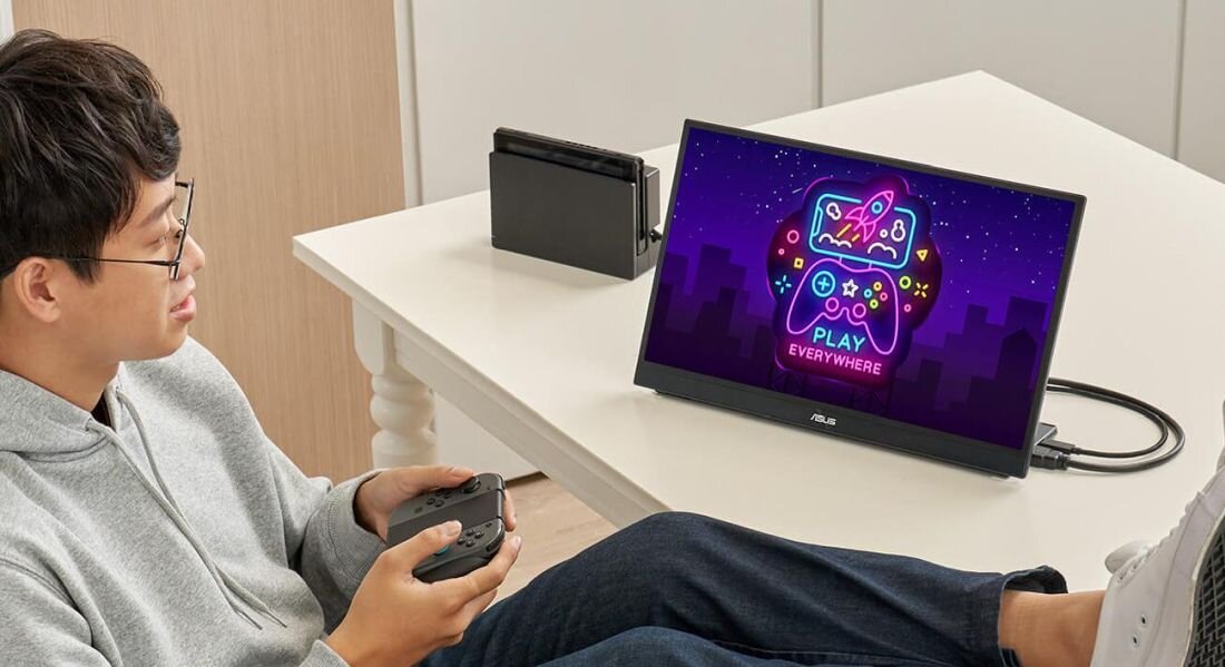 Osoba siedzi przy stole, trzymając kontroler do gier, patrząc na monitor z wyświetlonym kolorowym napisem \'PLAY EVERYWHERE\'. Obok monitora znajduje się konsola do gier.