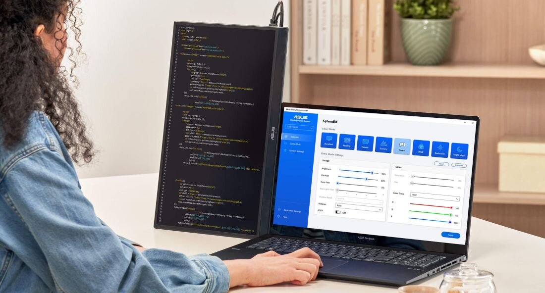 Osoba siedzi przy biurku z laptopem, na którym wyświetlone są ustawienia ekranu. Obok znajduje się monitor ASUS z pionowo wyświetlonym kodem programistycznym.