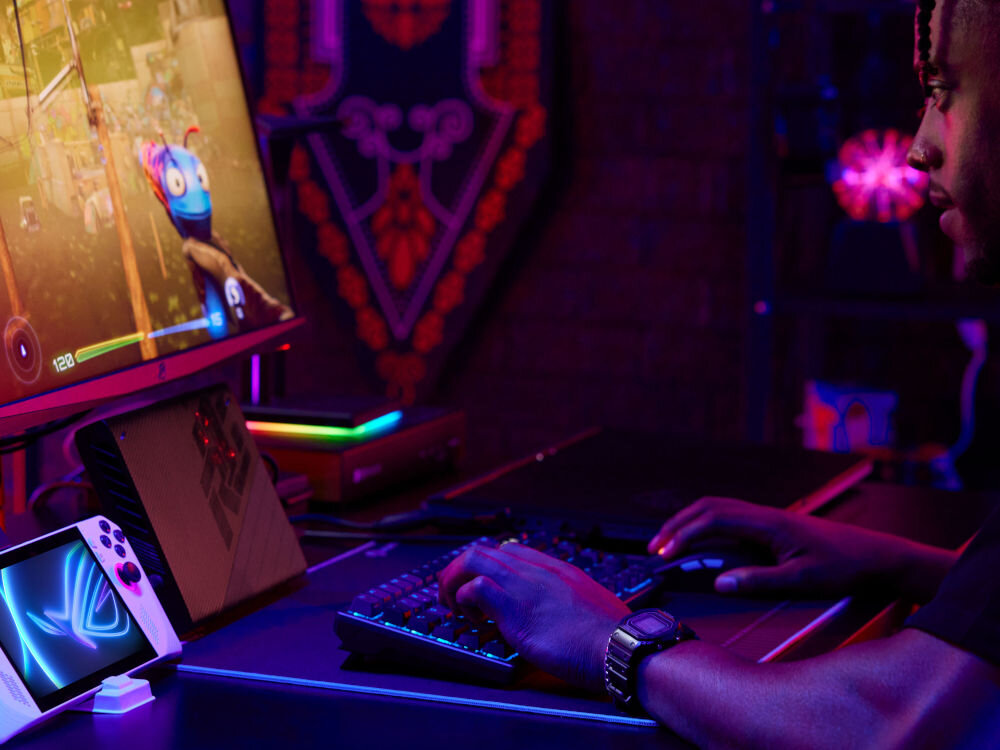 Osoba siedzi przy biurku z klawiaturą i monitorem, na którym wyświetlana jest gra. Obok znajduje się konsola z logo ASUS ROG, a w tle widoczne są kolorowe światła i dekoracje.