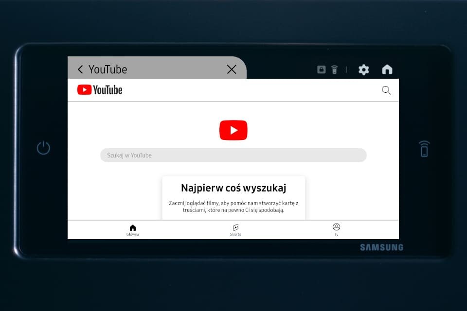 ekran youtube Ekran urządzenia wyświetla stronę YouTube z logo i polem wyszukiwania. Widoczny jest tekst: \'Najpierw coś wyszukaj Zaczni oglądać filmy, aby pomóc nam stworzyć kartę z treściami, które na pewno Ci się spodobają.\'