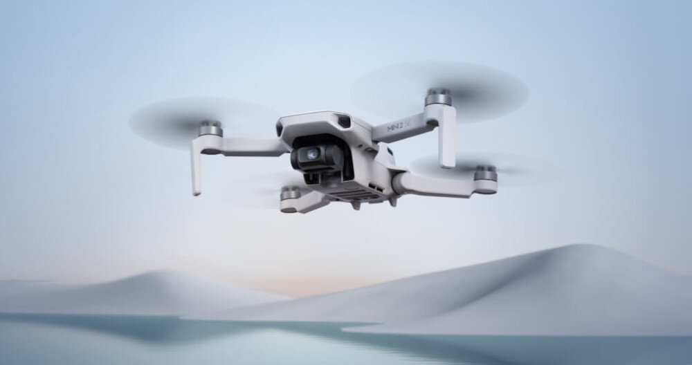 Na obrazie widoczny jest biały dron z czterema wirnikami, unoszący się w powietrzu na tle górskiego krajobrazu. Dron ma zamontowaną kamerę z przodu.