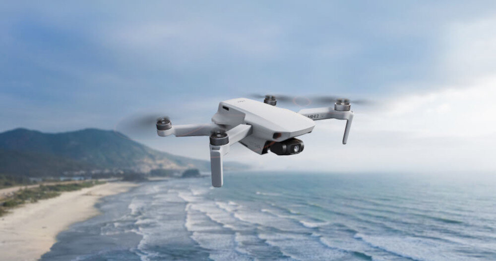 Dron unosi się nad wybrzeżem z widokiem na morze i góry w tle. Niebo jest częściowo zachmurzone.
