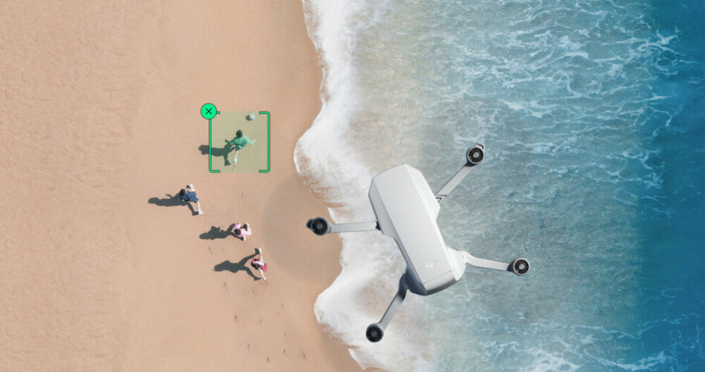 Na plaży widać trzy osoby, a nad nimi unosi się dron. Obok jednej z osób znajduje się zielony prostokąt z symbolem \'X\'.