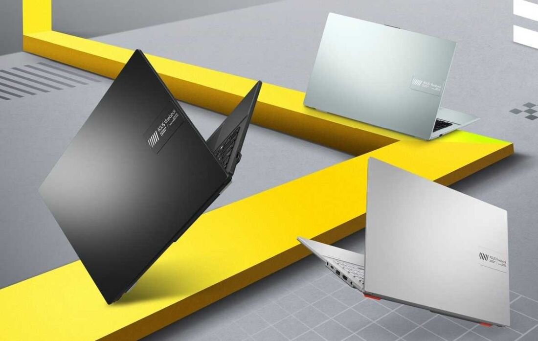 Trzy laptopy w różnych kolorach (czarny, srebrny, szary) ustawione na tle z żółtymi i szarymi elementami. Na każdym laptopie widoczne jest logo producenta z napisem \'ASUS VivoBook\'.