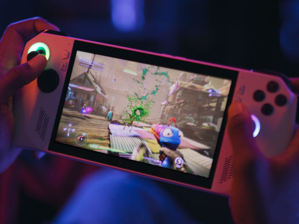 Osoba trzyma przenośną konsolę do gier z wyświetlaczem, na którym widoczna jest dynamiczna scena z gry. Konsola ma białą obudowę z przyciskami i joystickami po obu stronach ekranu.