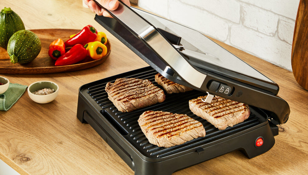Na drewnianym blacie stoi otwarty grill elektryczny z czterema kawałkami mięsa. Obok znajdują się miseczki z przyprawami i talerz z warzywami, takimi jak papryki i cukinie.