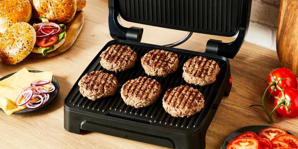 Na otwartym grillu elektrycznym znajduje się pięć grillowanych burgerów. Obok widoczne są składniki do burgerów, takie jak bułki, ser, cebula i pomidory.