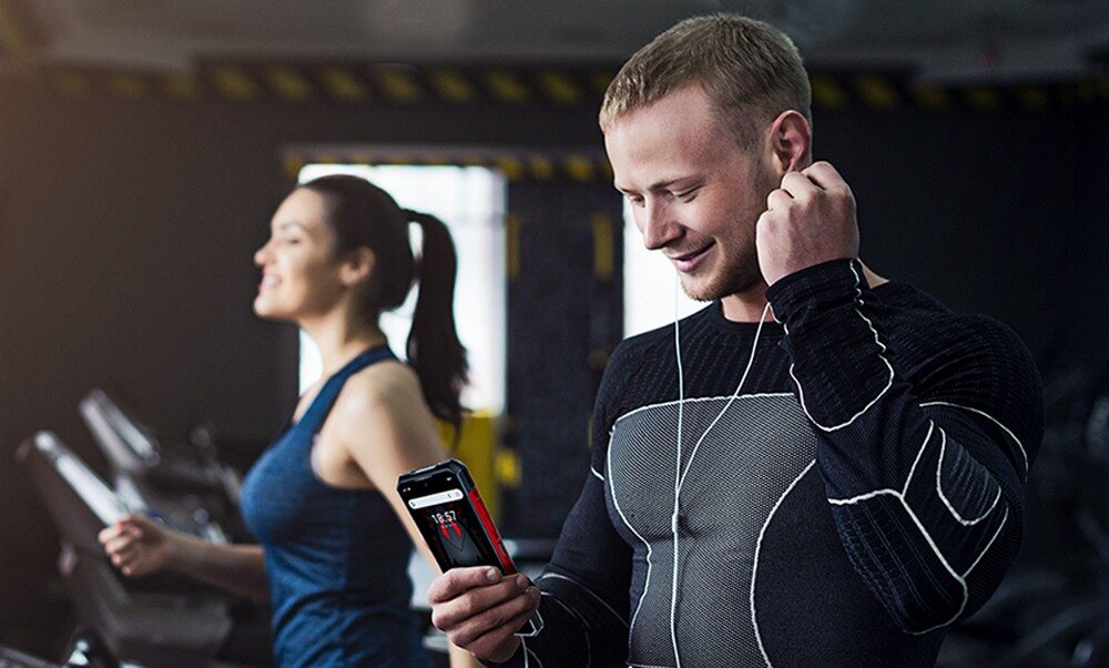 Mężczyzna w sportowym stroju trzyma smartfon, na którym widoczny jest ekran z aplikacją. W tle znajduje się kobieta ćwicząca na bieżni.