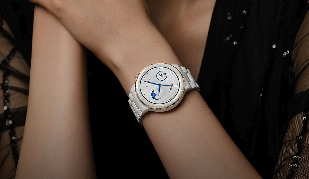 Osoba nosi elegancki smartwatch z białą bransoletą i okrągłą tarczą, na której widoczne są wskazówki i subtelne detale graficzne. Tło jest ciemne, a ubranie osoby ma delikatne, przezroczyste elementy.
