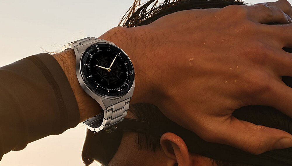 Osoba nosi srebrny smartwatch z czarną tarczą na nadgarstku. W tle widać rozmyty krajobraz o zachodzie słońca.