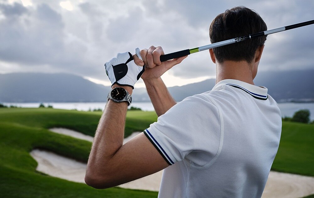 Osoba w białej koszulce trzyma kij golfowy na tle pola golfowego. Na nadgarstku widoczny jest smartwatch z okrągłą tarczą.