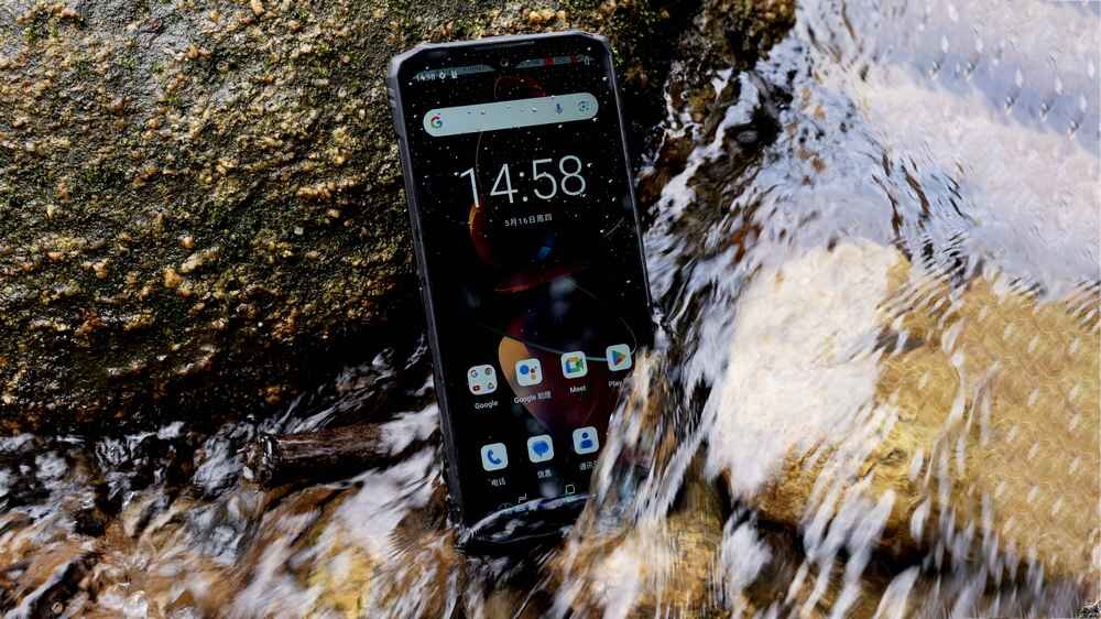 Smartfon leży w płytkiej wodzie obok kamienia, na ekranie widoczna jest godzina 14:58 oraz kilka ikon aplikacji. Ekran jest włączony, a na powierzchni urządzenia widać krople wody.
