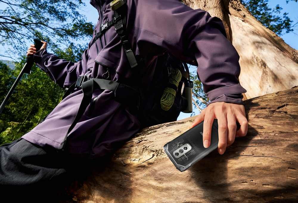 Osoba w fioletowej kurtce wspiera się na dużym pniu drzewa, trzymając w ręce czarny smartfon. W tle widać zielone liście drzew i niebo.
