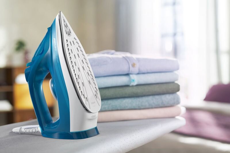 Na desce do prasowania stoi żelazko w kolorze niebiesko-białym z widocznym logo \'PHILIPS\'. Obok znajduje się stos złożonych ubrań w różnych kolorach.