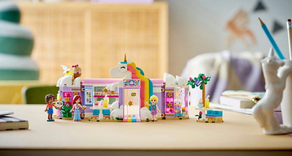 Zestaw LEGO przedstawiający kolorową kawiarnię z motywem jednorożca, z kilkoma figurkami postaci i dekoracjami w tle. Na pierwszym planie widoczne są różnokolorowe elementy konstrukcyjne oraz detale takie jak rośliny i meble.