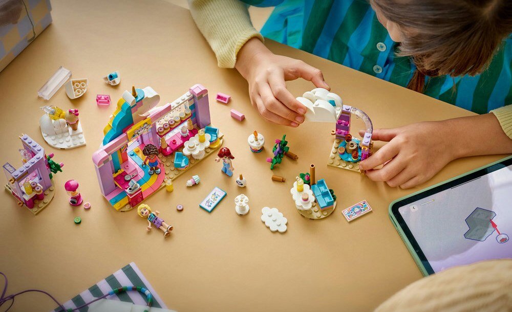 Na stole znajdują się kolorowe elementy zestawu LEGO, w tym miniaturowe postacie i akcesoria. Dziecko bawi się klockami, a obok leży tablet z wyświetlonym obrazem.