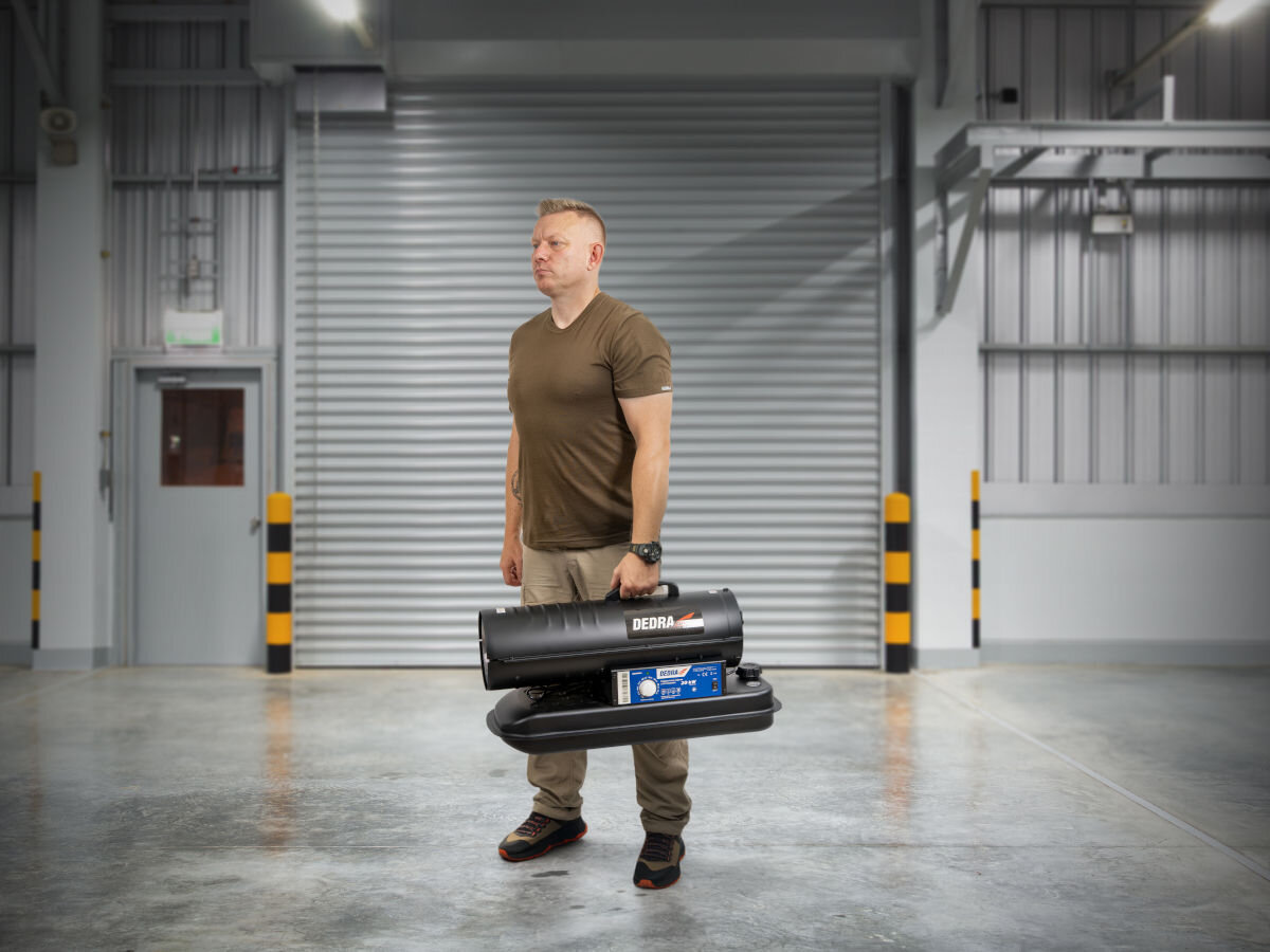 Nagrzewnica olejowa DEDRA DED9950A, uchwyt, metal, stabilność, wskaźnik, Osoba trzyma nagrzewnicę Dedra w hali; uchwyt umożliwia wygodne przenoszenie urządzenia.