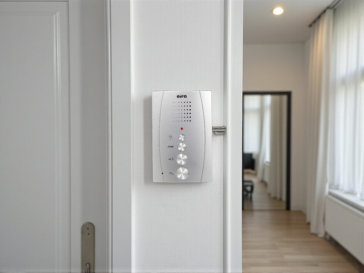 Unifon EURA ADA-13A3 Srebrny, komunikacja, ochrona, bezpieczeństwo, wejścia, Unifon EURA ADA na ścianie naprzeciw drzwi wejściowych, w tle korytarz budynku.