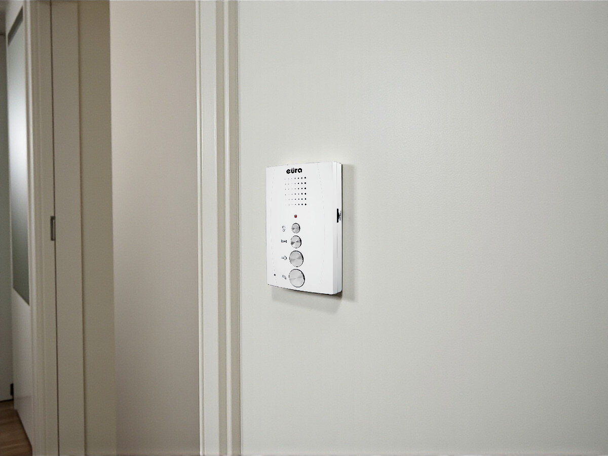 Unifon EURA ADA-11A3 Biały, funkcjonalność, bezpieczeństwo, czteroprzewodowy, montaż, Unifon EURA ADA-11A3 na ścianie przed drzwiami, jasne tło, boczny widok urządzenia w korytarzu.