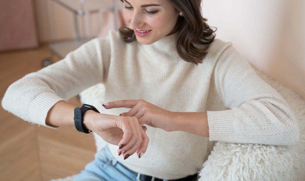 Osoba siedzi na fotelu i patrzy na szary smartwatch na swoim nadgarstku. Ubrana jest w jasny sweter i dżinsy.