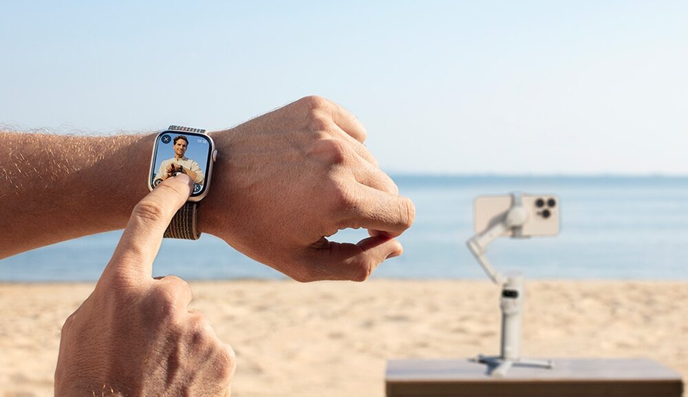 Osoba dotyka ekranu smartwatcha, na którym widoczna jest inna osoba. W tle na plaży stoi smartfon zamocowany na gimbalu.