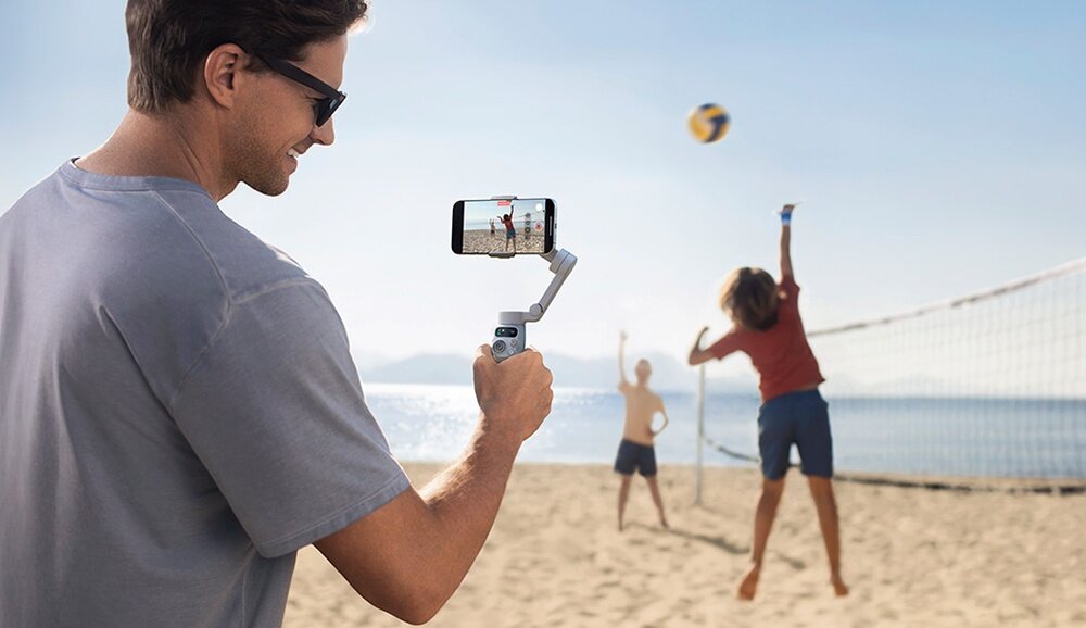 Osoba trzyma gimbal z zamocowanym smartfonem, na ekranie którego widać scenę z gry w siatkówkę plażową. W tle znajduje się plaża, siatka do siatkówki i dwie osoby grające w piłkę.