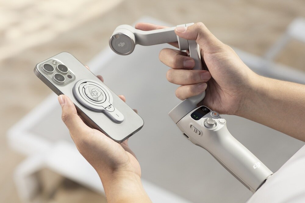 Osoba trzyma szary gimbal DJI Osmo Mobile 7 oraz smartfon z zamontowanym uchwytem magnetycznym z logo DJI. W tle widać rozmyty piasek i fragment białego mebla.