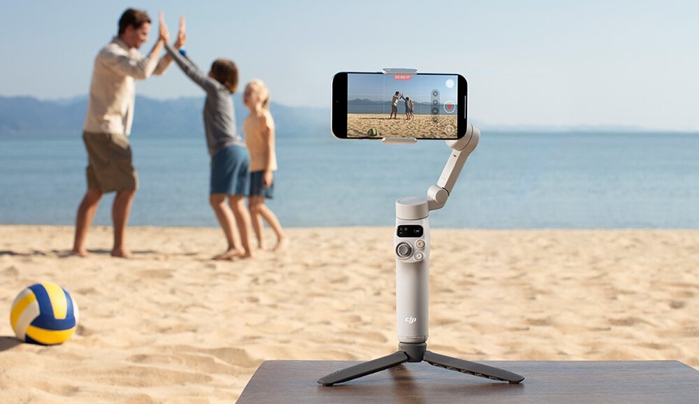 Na plaży stoi szary gimbal z zamocowanym smartfonem, na którym widać nagrywany obraz dwóch osób bawiących się na piasku. Obok leży piłka plażowa, a w tle rozciąga się morze.