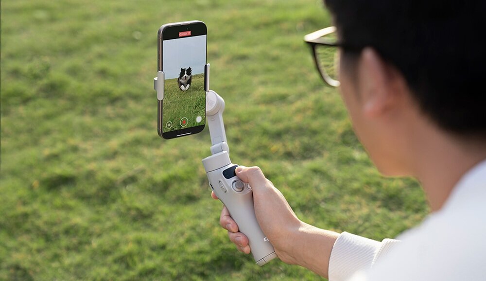 Osoba trzyma szary gimbal, na którym zamocowany jest smartfon. Na ekranie telefonu widać psa na trawie.