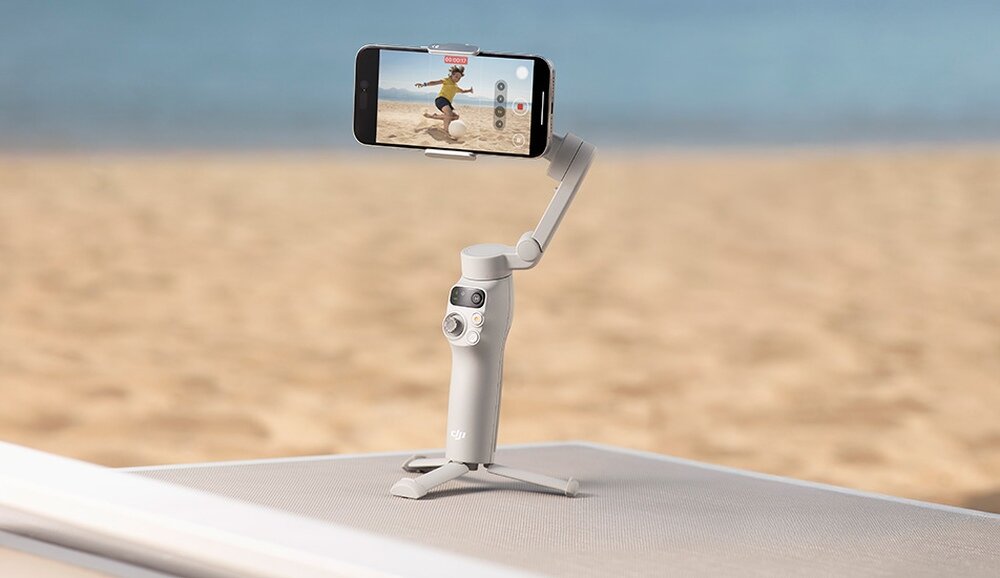 Szary gimbal z zamocowanym smartfonem stoi na powierzchni, w tle widać plażę i morze. Na ekranie smartfona widoczny jest obraz osoby biegnącej po piasku.