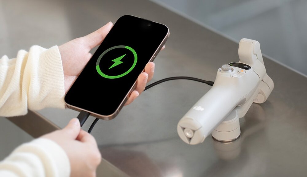Osoba trzyma smartfon z wyświetlonym symbolem ładowania, podłączony do szarego gimbala z logo DJI. Gimbal leży na metalowej powierzchni.