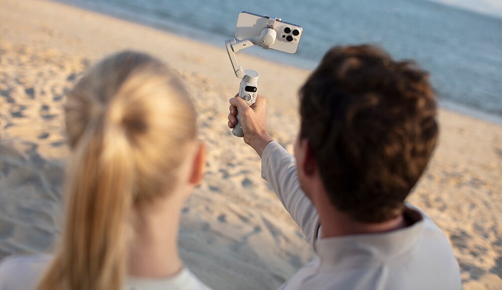 Osoba trzyma gimbal z zamocowanym smartfonem na plaży, w tle widać morze.
