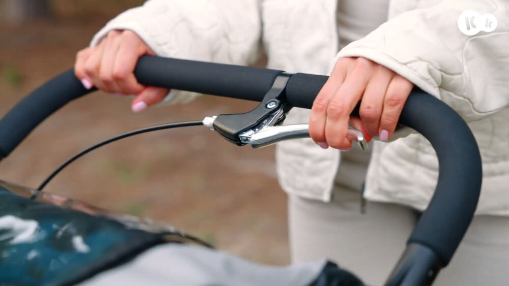 Osoba trzyma uchwyt przyczepki rowerowej z czarną, miękką rączką i srebrnym mechanizmem hamulca. W górnym rogu widoczne jest logo \'Kinderkraft\'.