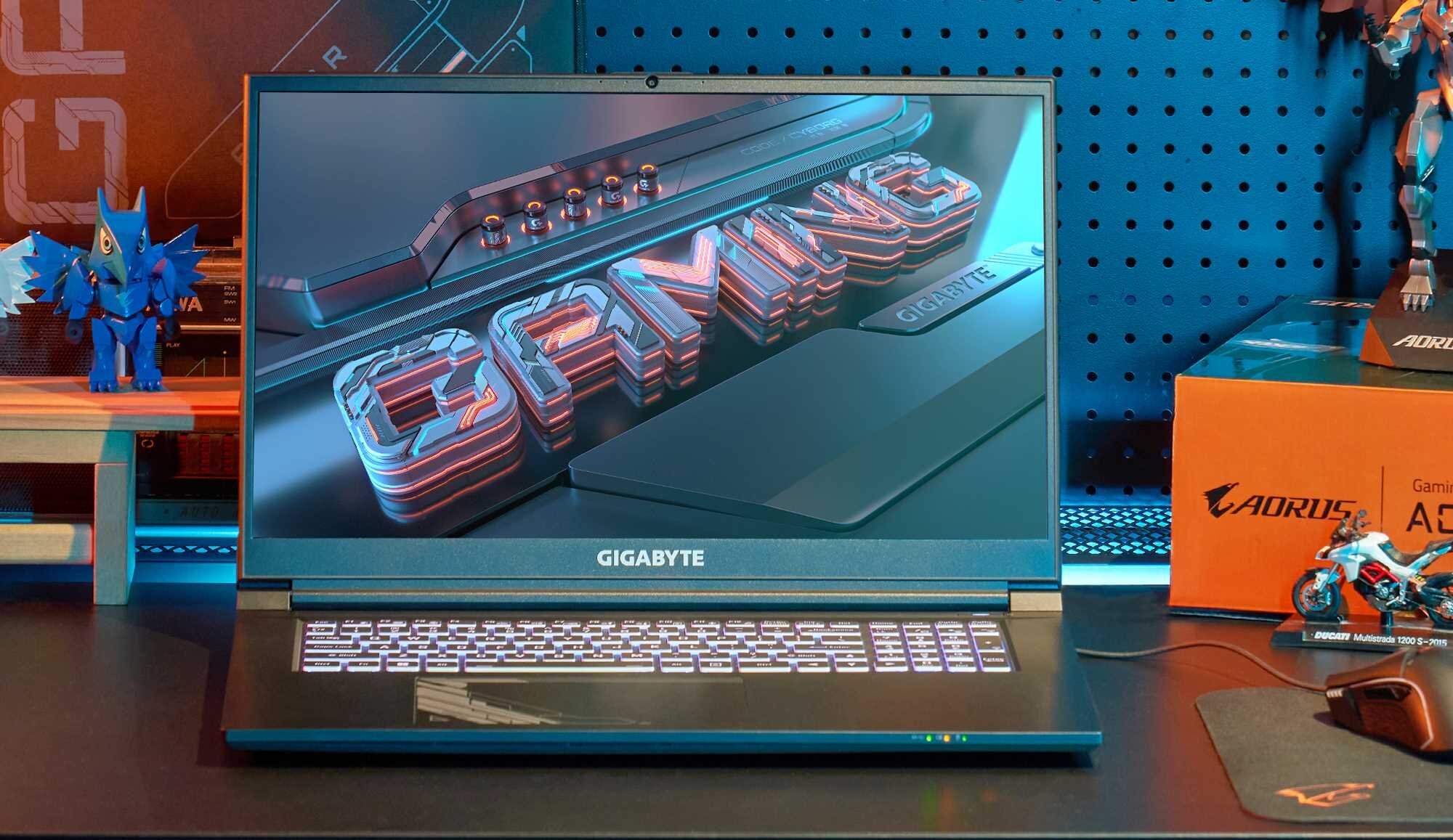 Laptop GIGABYTE z wyświetlaczem pokazującym napis \'GAMING\' w stylu 3D. Na biurku obok laptopa znajdują się figurki i pudełko z logo AORUS.