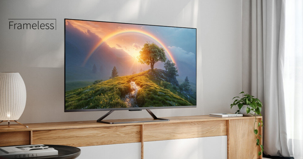 Telewizor z cienką ramką wyświetla krajobraz z tęczą nad zielonym wzgórzem. Obok telewizora znajduje się lampa i roślina, a na ekranie widoczny jest napis \'Frameless\'.