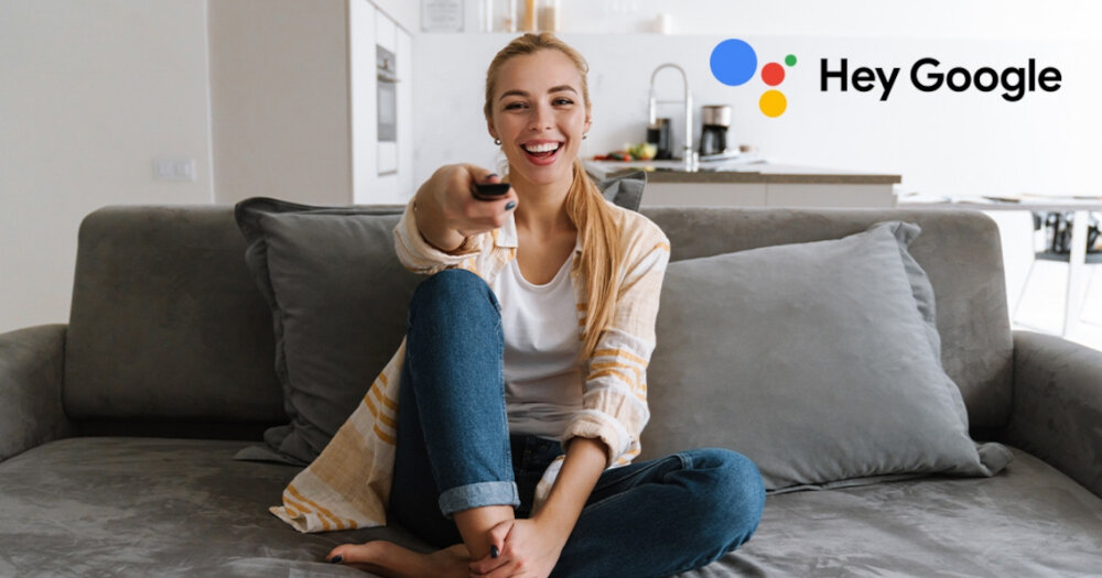 Osoba siedzi na kanapie z pilotem w ręku, uśmiechając się. W tle widoczne jest logo Google z napisem \'Hey Google\'.