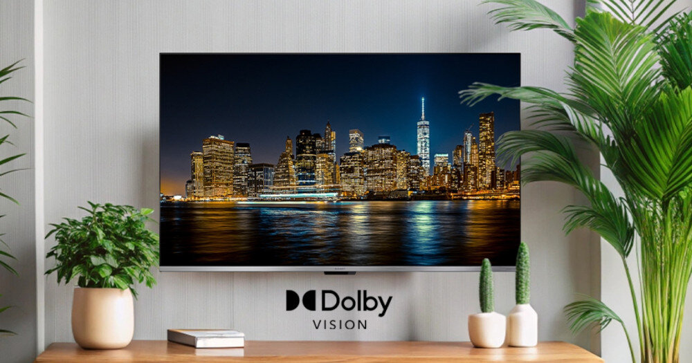 Telewizor wyświetla nocny widok na miasto z odbiciem świateł w wodzie. Pod ekranem widoczne jest logo \'Dolby Vision\', a wokół znajdują się rośliny doniczkowe i książka.