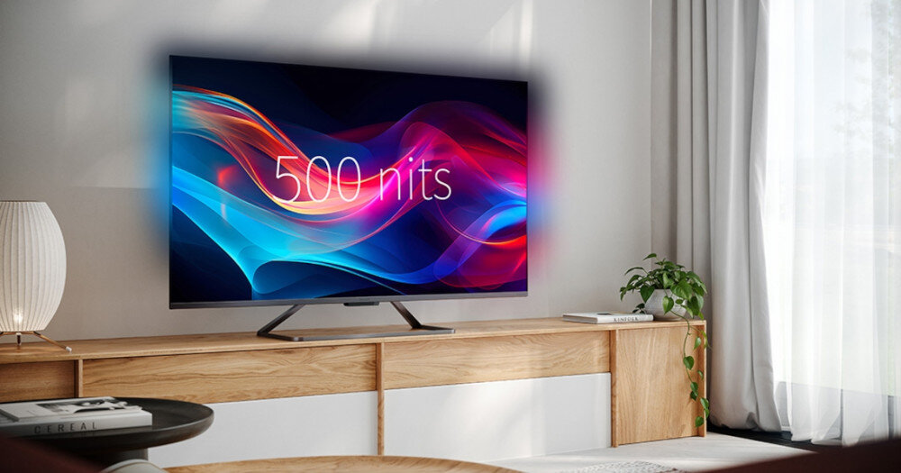 Telewizor wyświetla kolorową grafikę z napisem \'500 nits\'. Stoi na drewnianej szafce w jasnym, nowoczesnym wnętrzu.