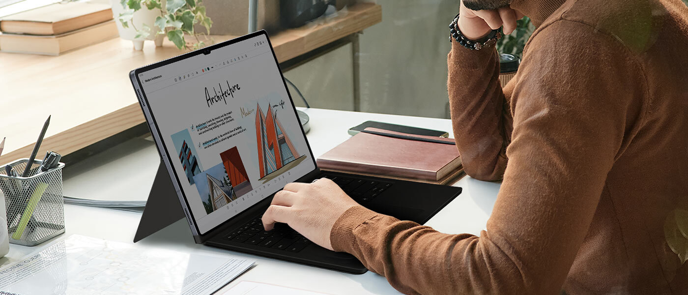 Mężczyzna korzysta z Galaxy Tab S10, trzymając dłoń na etui Book Cover Keyboard. Osoba siedzi przy biurku, używając tabletu z klawiaturą w etui. Na ekranie widoczna jest strona internetowa z napisem \'Architecture\' i obrazkami budynków.