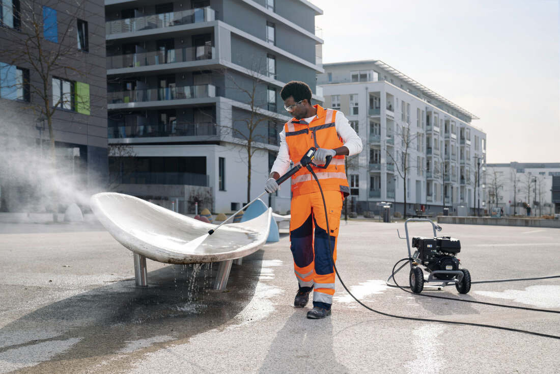 Osoba w pomarańczowym stroju roboczym używa myjki ciśnieniowej do czyszczenia dużego, metalowego obiektu na zewnątrz. W tle widoczne są nowoczesne budynki mieszkalne.