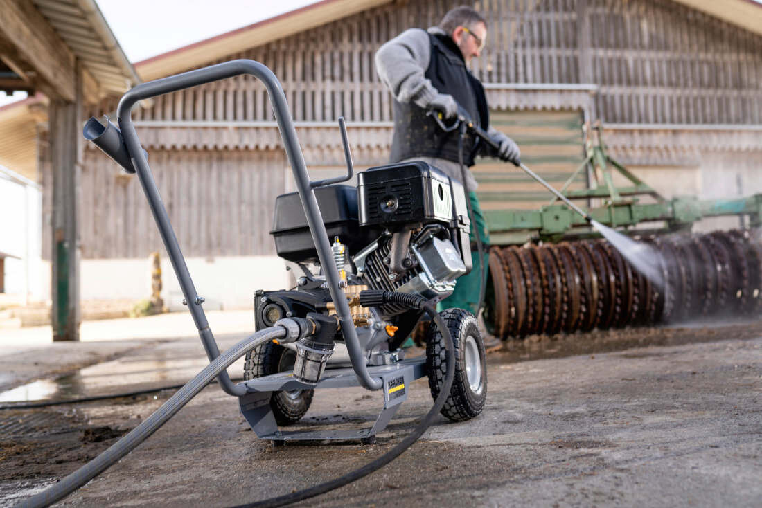 Na obrazie widać myjkę ciśnieniową na kółkach z widocznym logo KARCHER. W tle osoba używa myjki do czyszczenia dużego, metalowego urządzenia.