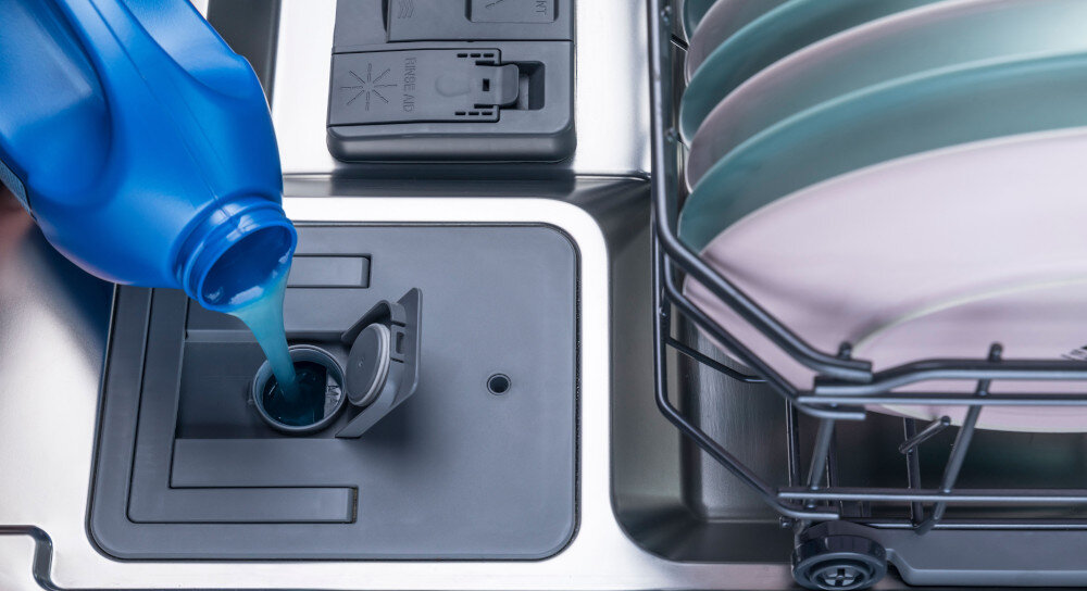 Osoba nalewa płyn do pojemnika w zmywarce, obok widoczne są ułożone talerze w koszu. Na pokrywie pojemnika widoczny jest symbol płatka śniegu i napis \'rinse aid\'.