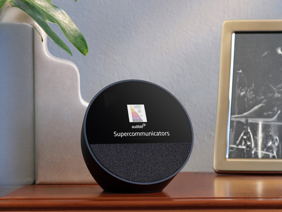 Czarny głośnik z wyświetlaczem, na którym widnieje napis \'audible Supercommunicators\'. Obok znajduje się roślina i ramka ze zdjęciem.