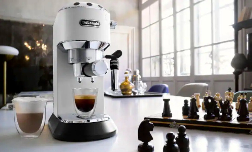 Na blacie stoi biały ekspres do kawy z logo \'DeLonghi\', obok filiżanka z kawą i szklanka z mlecznym napojem. W tle widać szachy i duże okna wpuszczające światło do pomieszczenia.