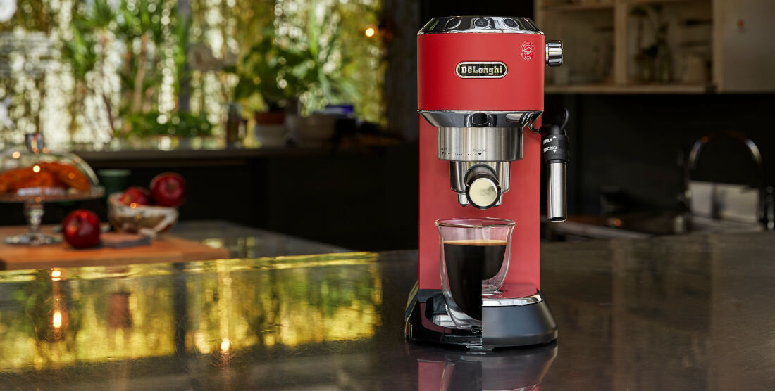 Czerwony ekspres do kawy z logo \'DeLonghi\' stoi na blacie, z filiżanką kawy pod dyszą. W tle widoczna jest kuchnia z roślinami i owocami na stole.