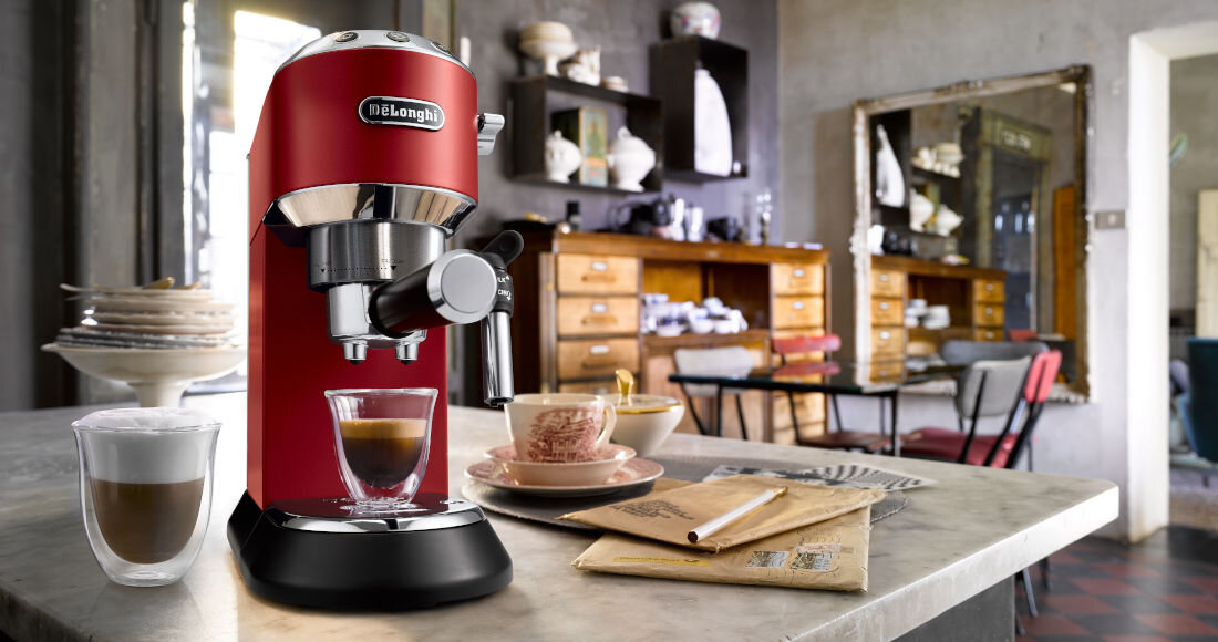 Na blacie kuchennym stoi czerwony ekspres do kawy z logo \'DeLonghi\', obok filiżanka z kawą i szklanka z mlekiem. W tle widoczna jest kuchnia z drewnianymi szafkami i półkami z naczyniami.