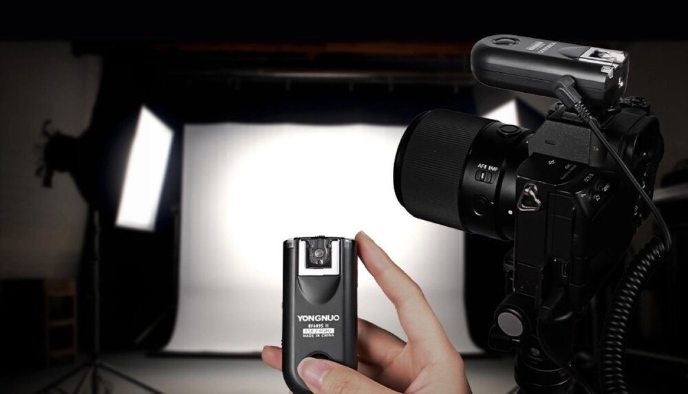 Jeden z wyzwalaczy radiowych Yongnuo RF603C trzymany w dłoni, obok aparat z zamontowanym wyzwalaczem, w tle studio